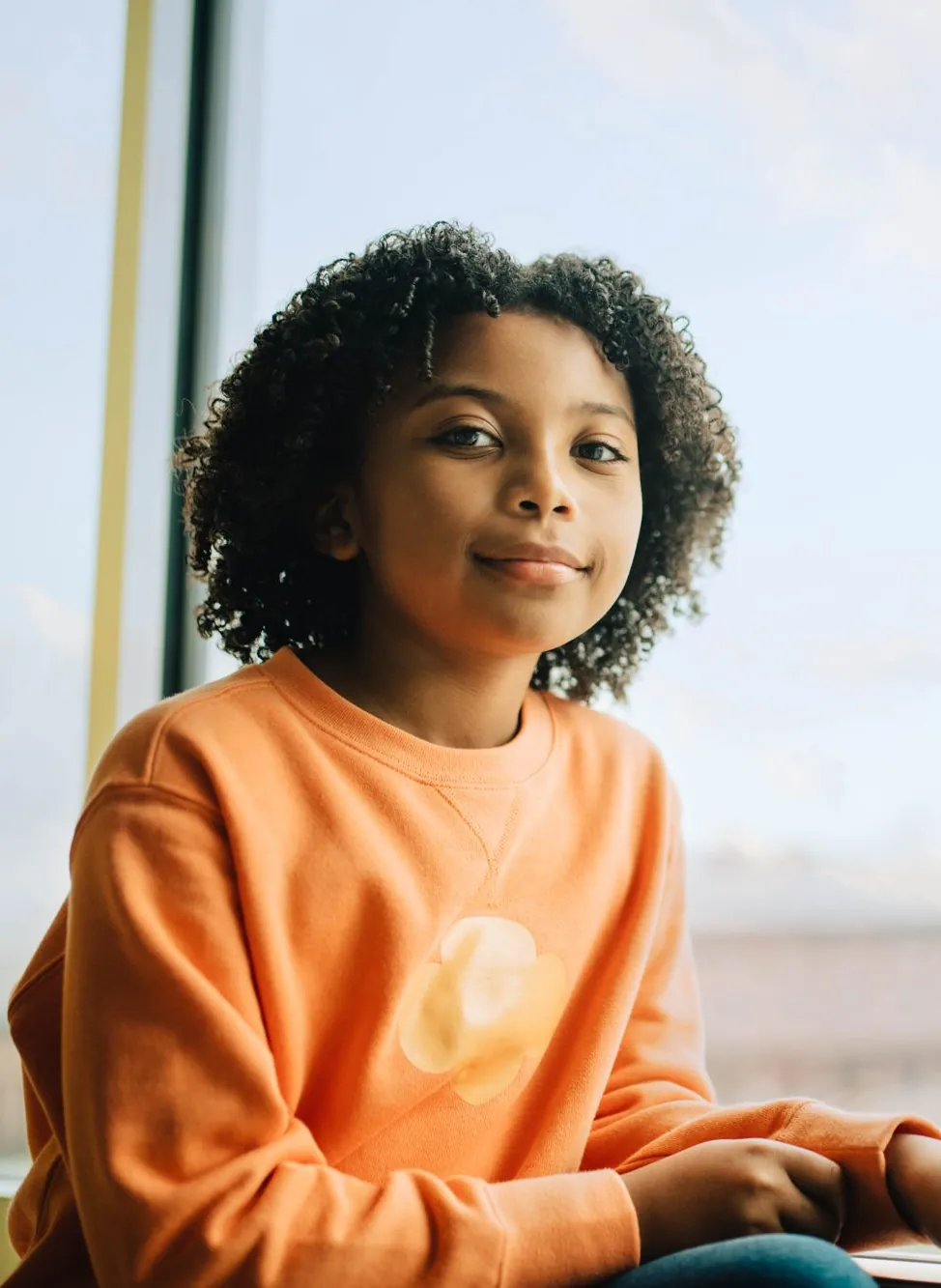 A smiling Girl Scout with curly hair in an orange sweatshirt sits by a sunny window
